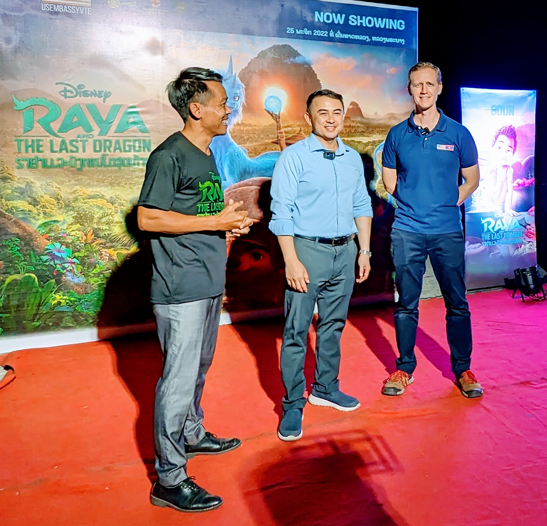 Steve Arounsack stands with two other men in front of a movie poster for "Raya and the Last Dragon."