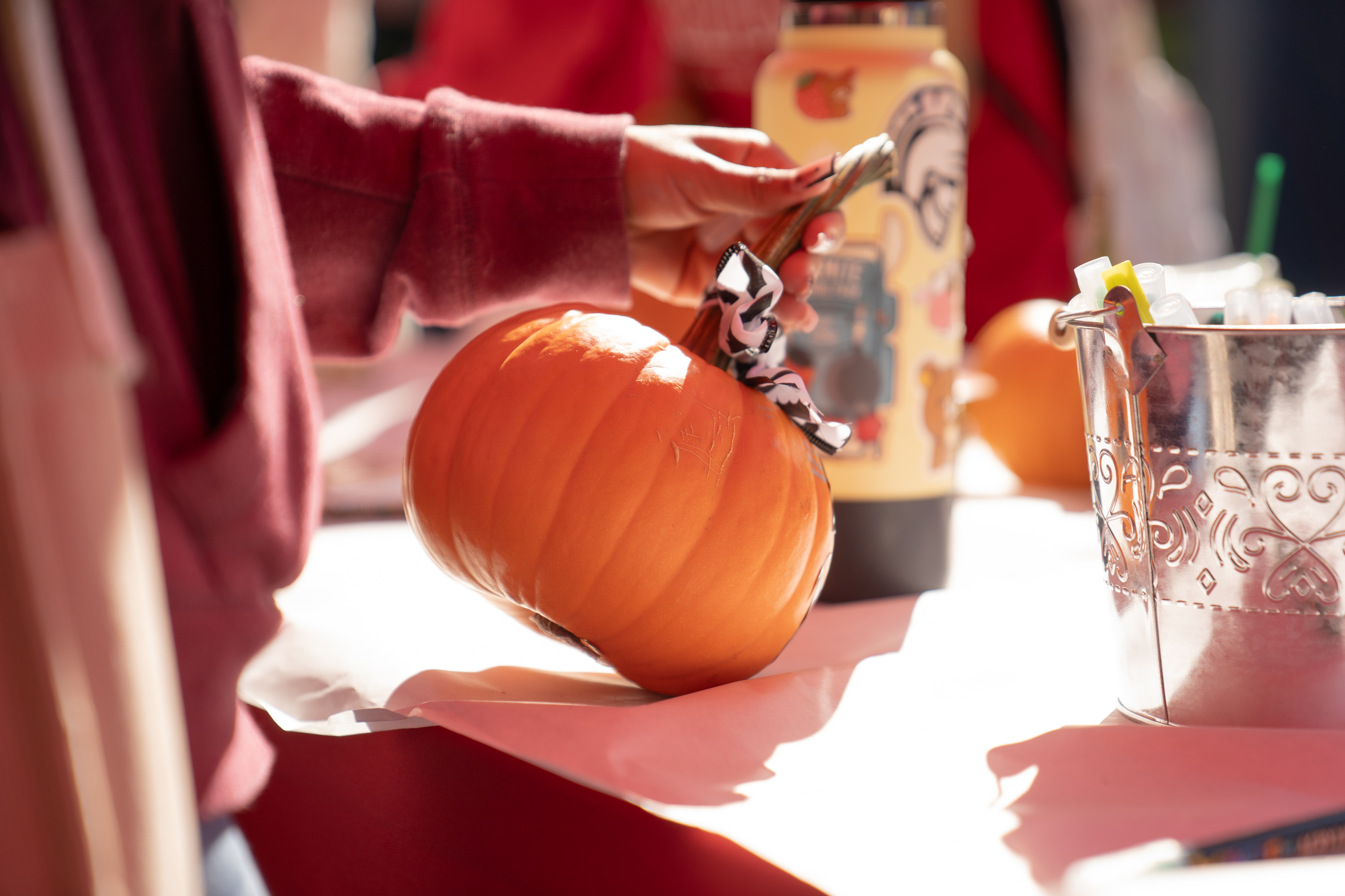 A person holds a small pumpkin on a table next to a decorated water bottle and a metal bucket.
