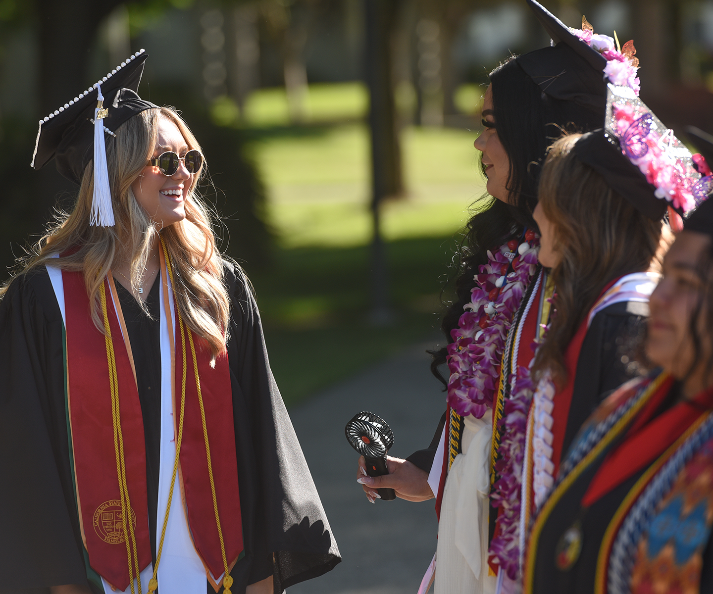 Graduates in caps and gowns celebrate outdoors, smiling and talking. They wear leis and stoles, conveying joy and accomplishment in bright sunlight.