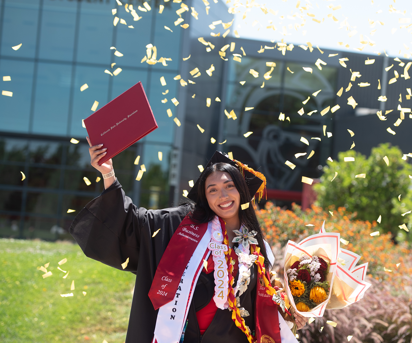 A joyful graduate in cap and gown celebrates with a diploma and bouquet, surrounded by confetti against a sunny backdrop.