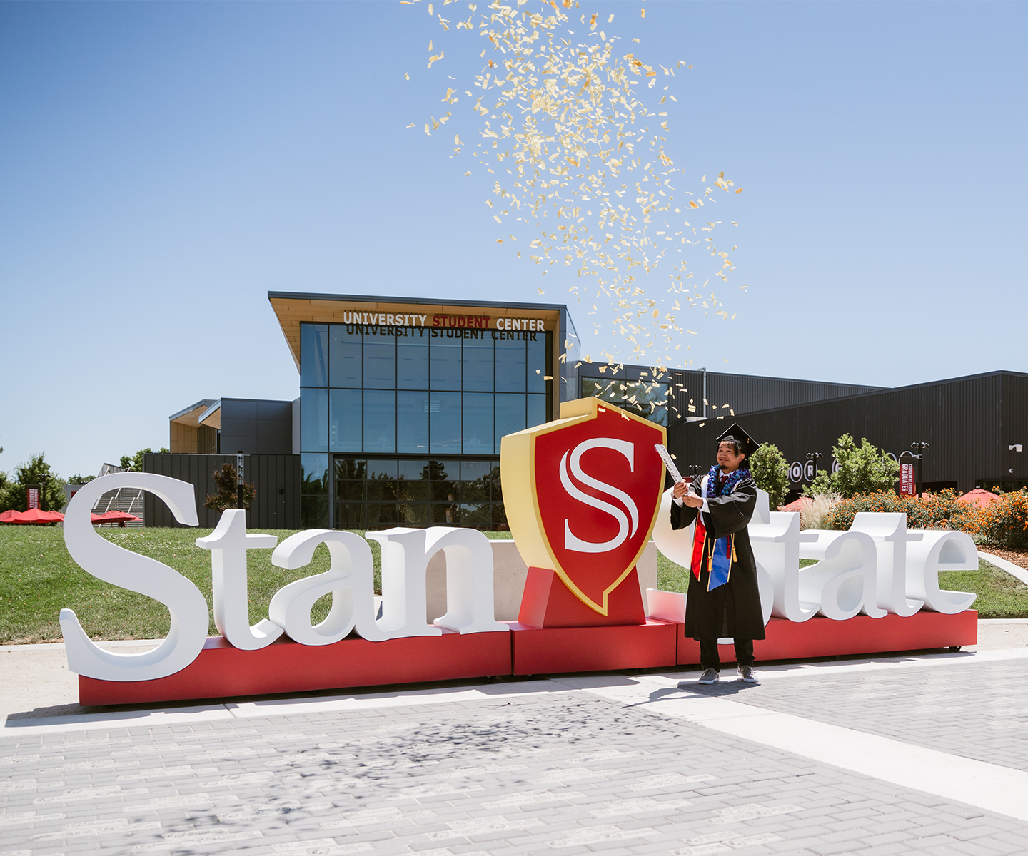 Graduate in cap and gown celebrates with confetti by a "Stan State" sign, in front of the University Student Center, under a clear blue sky.