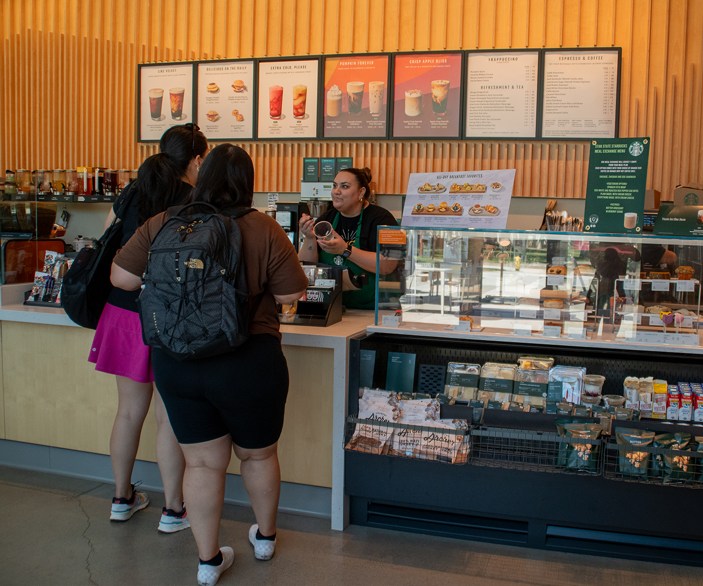 Barista engaging with two customers at a coffee shop counter, featuring a menu board above and a display of pastries and snacks below, creating a friendly and inviting atmosphere.
