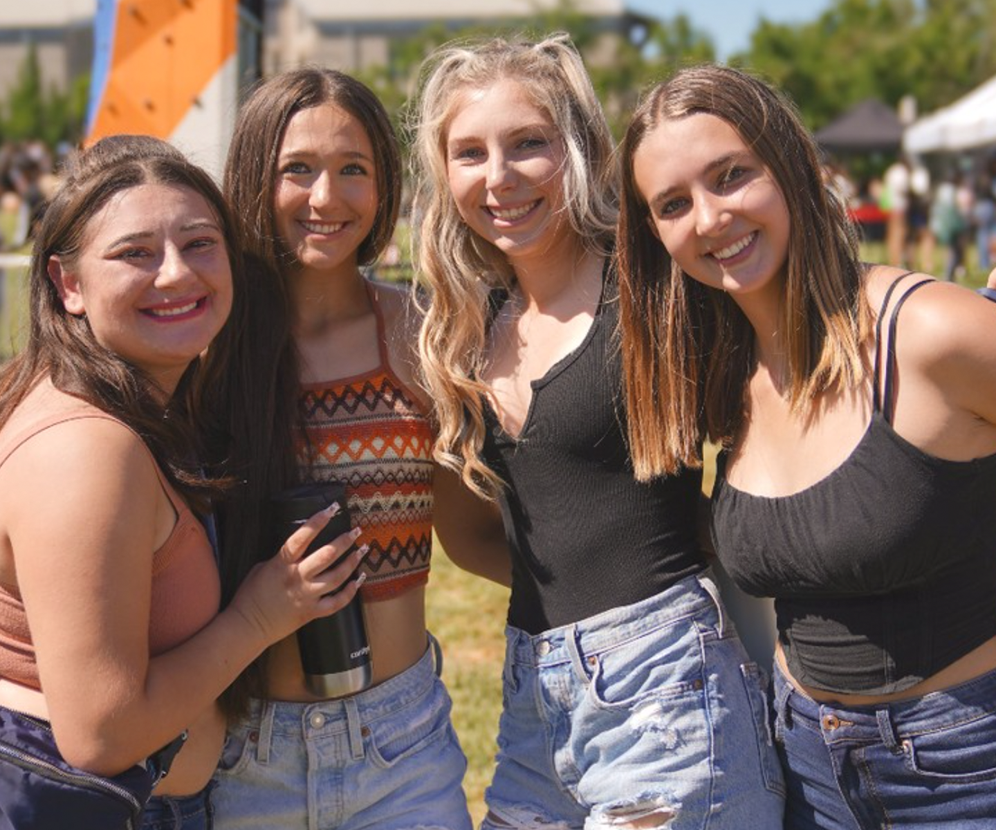 Four young women stand closely in a sunny outdoor setting, all smiling warmly. They wear casual summer outfits, conveying a joyful and carefree mood.