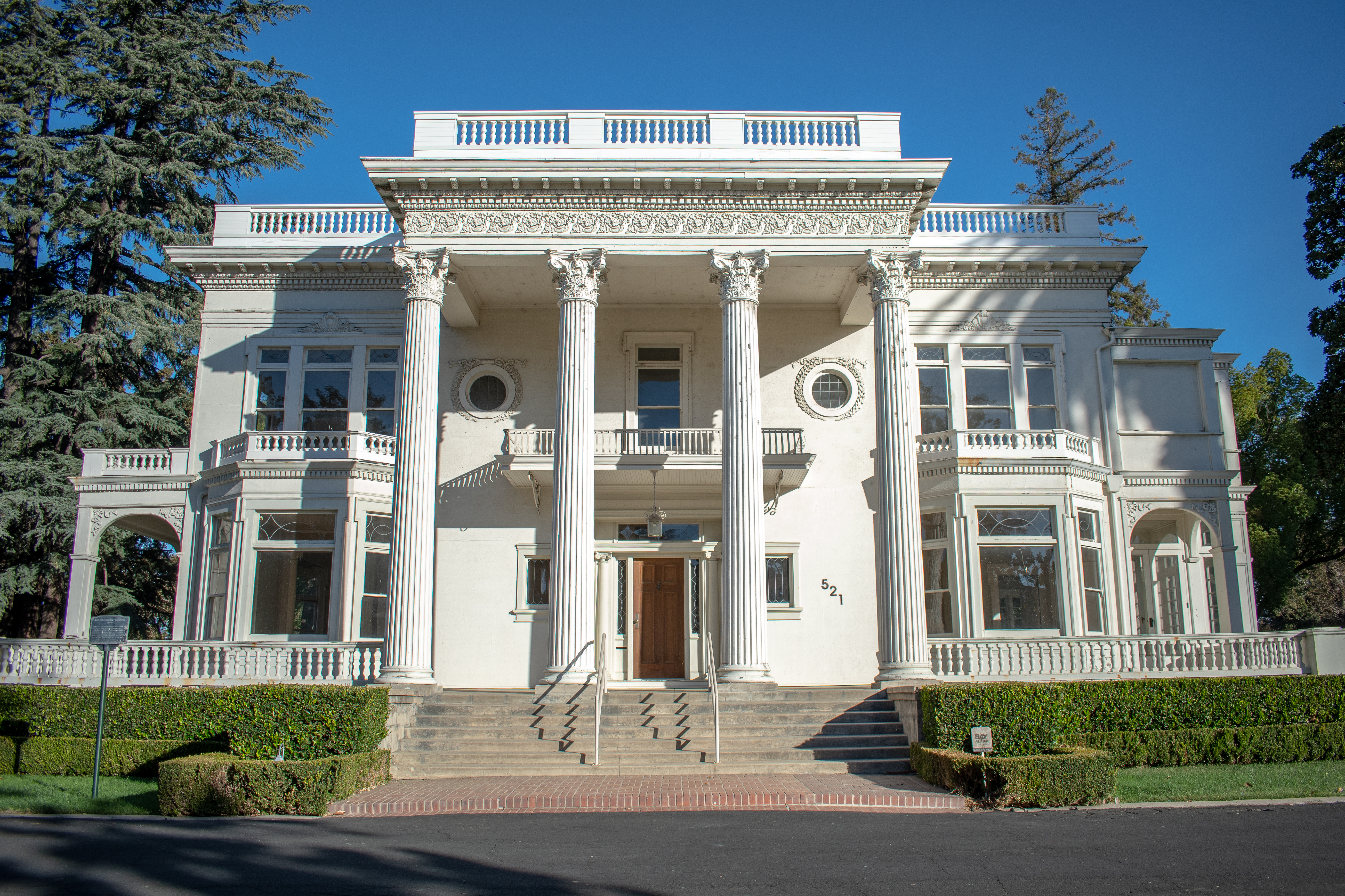 Historic white mansion with classical columns, ornate detailing, and a grand entrance. Surrounding are trimmed hedges and tall trees under a clear blue sky.