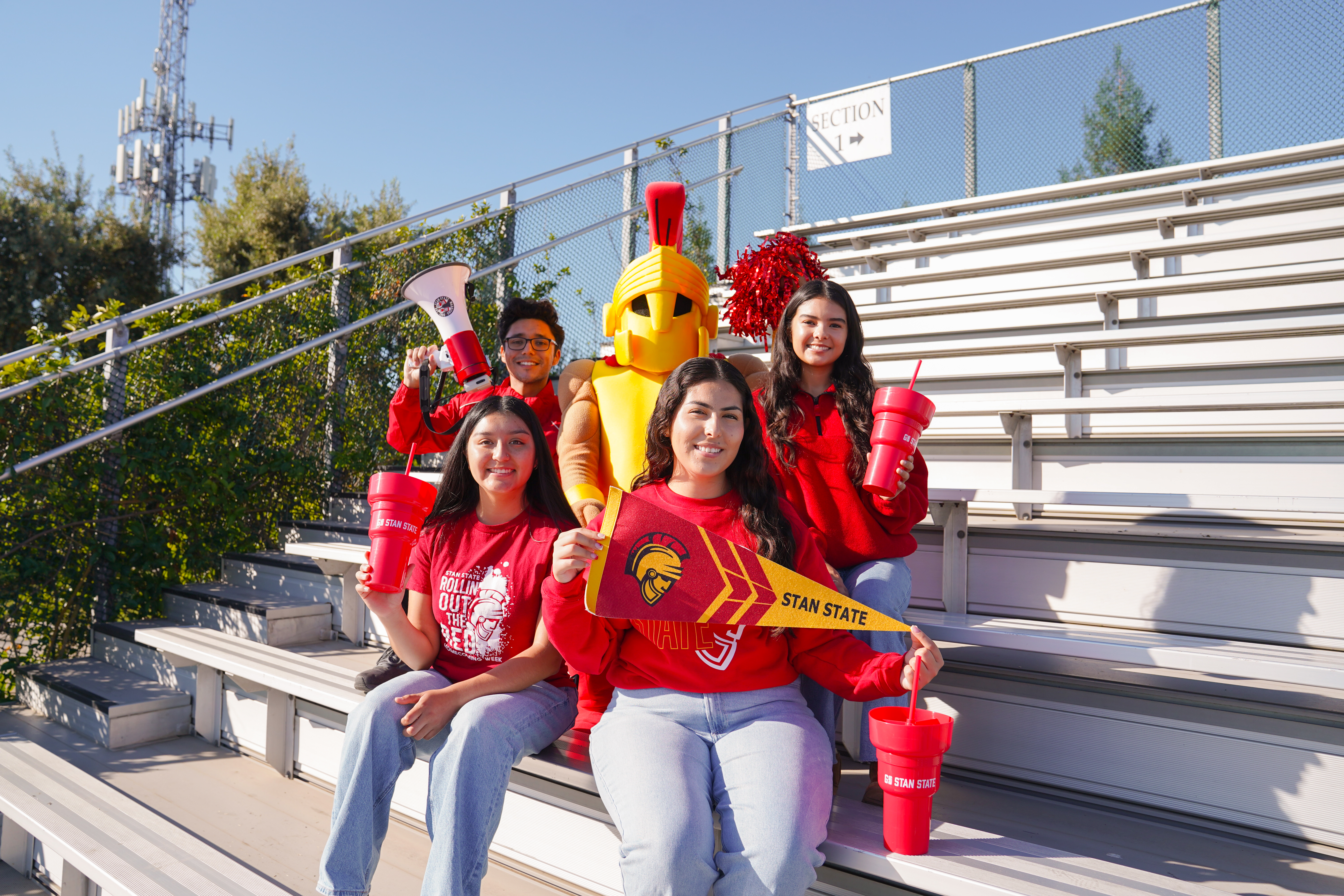 4 students sitting on the bleachers with Titus holding Stan State spirit merch