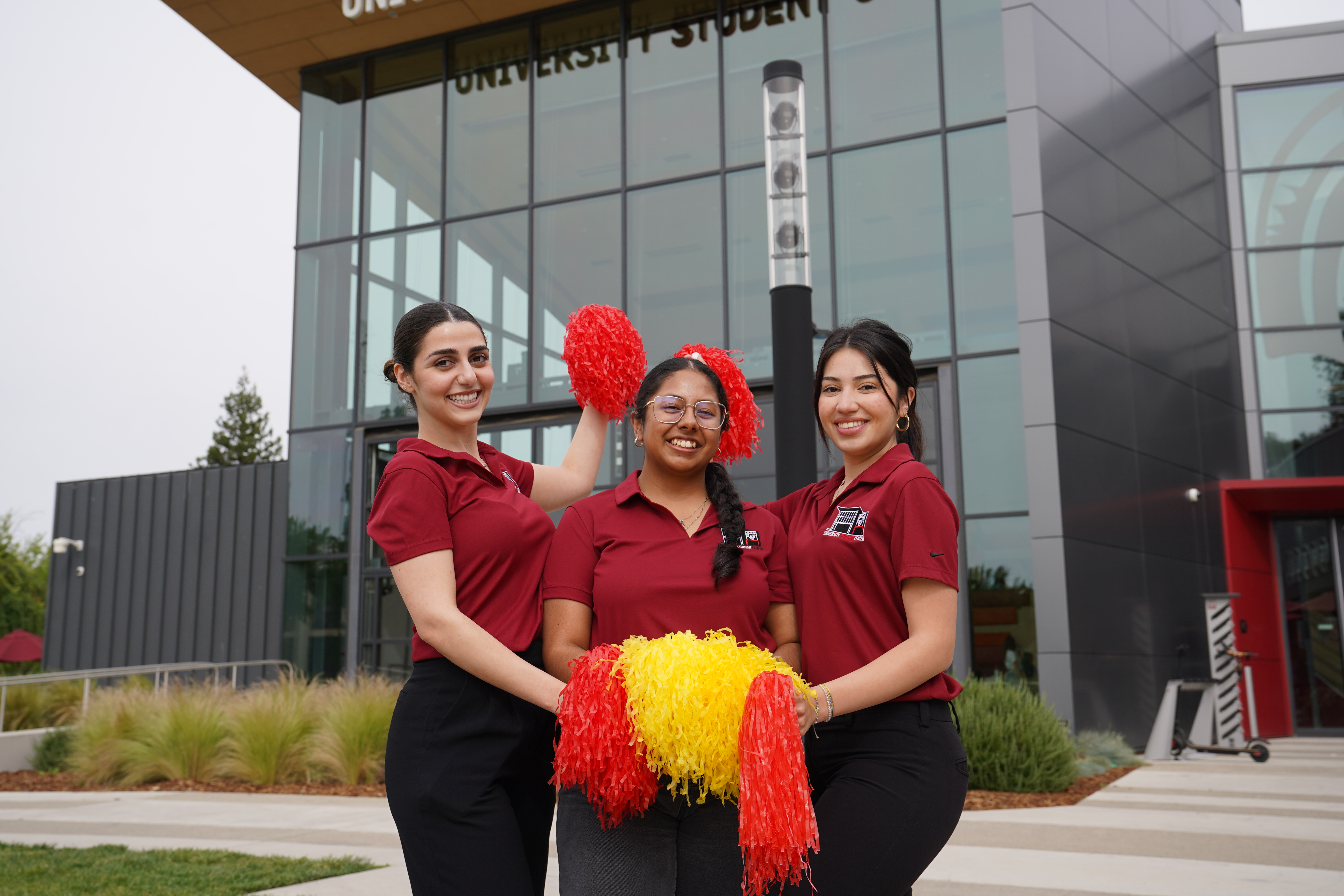 3 students holding pom poms posed in front of the University Student Center