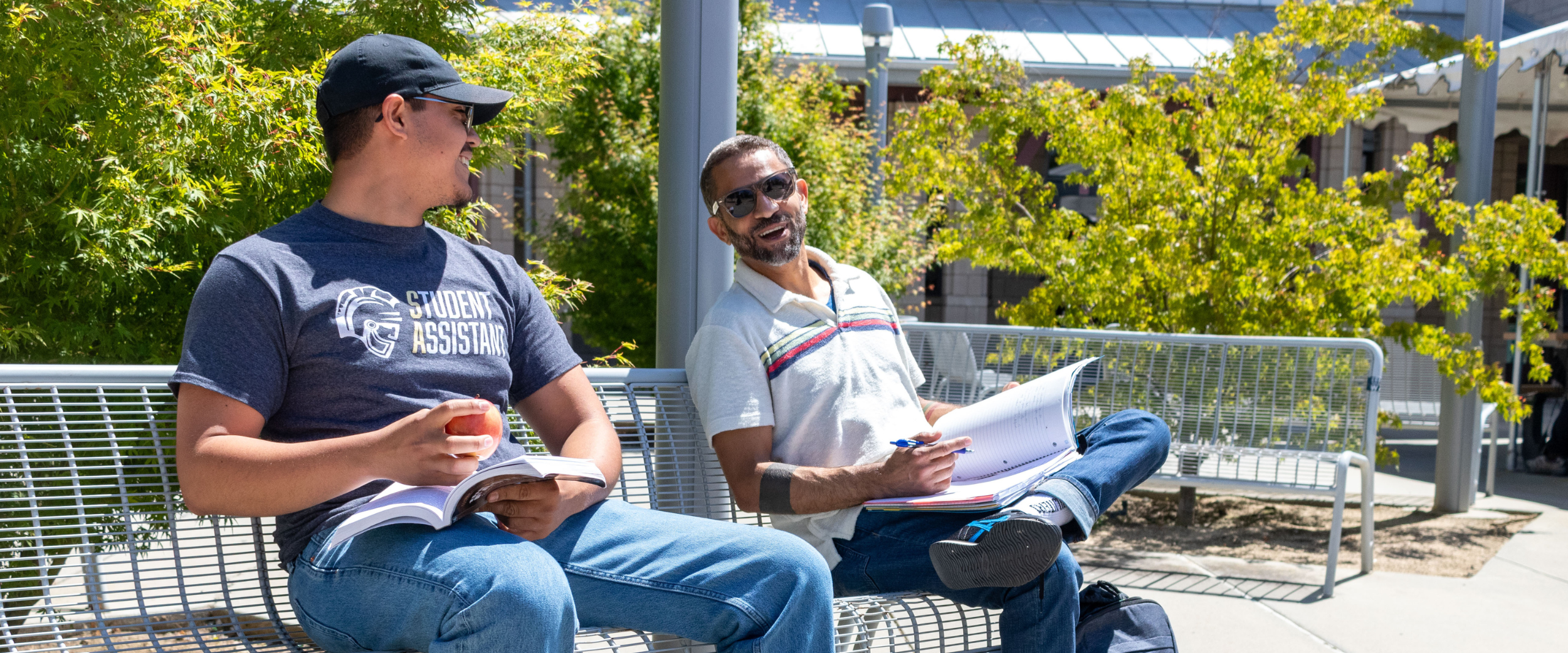 Two Stan State transfer students sitting on an outdoor bench, smiling and studying together with notebooks open and backpacks at their feet on a sunny day