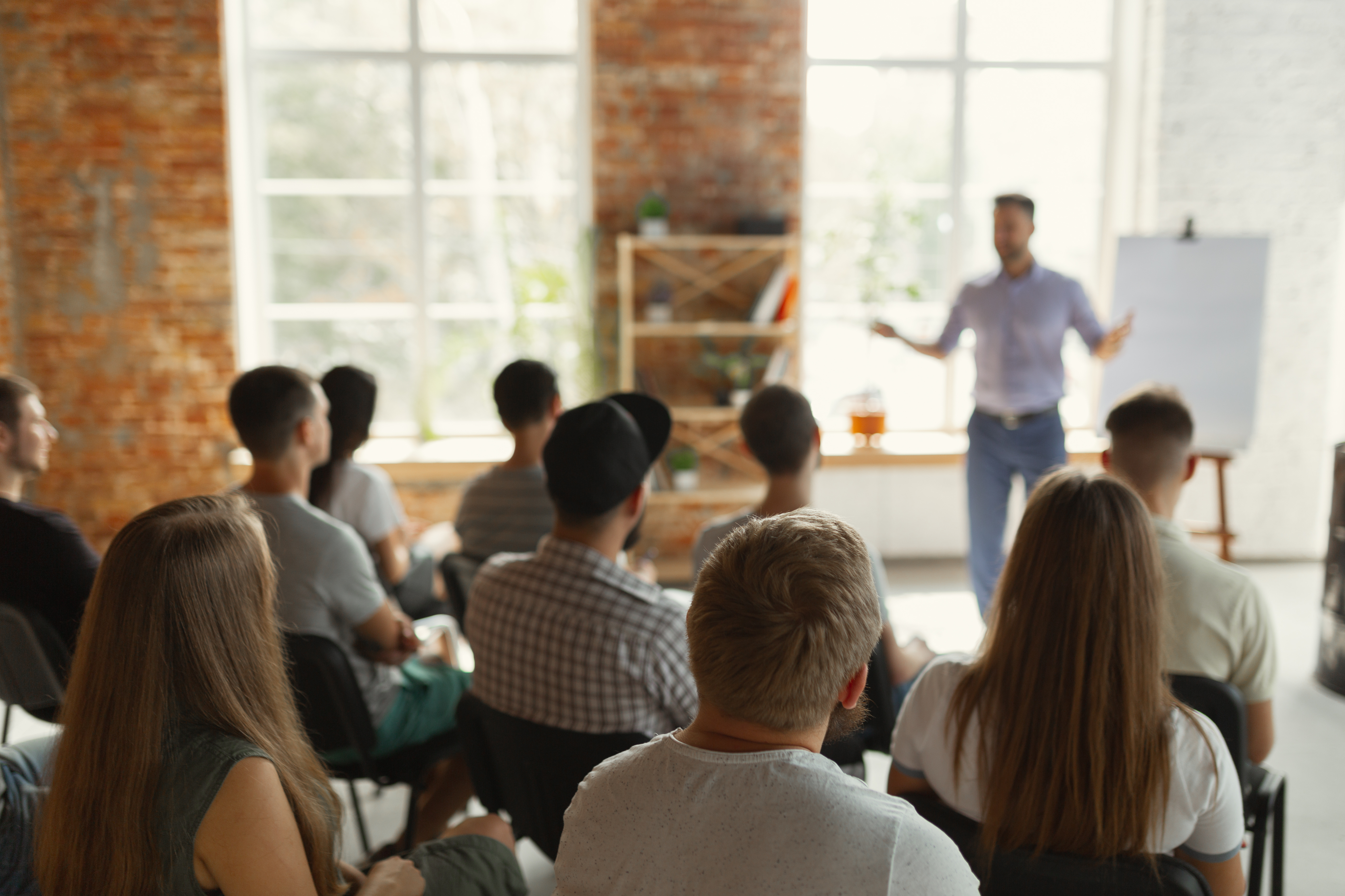 Audience members seated in a bright room listen to a speaker presenting at the front near a whiteboard and large windows.