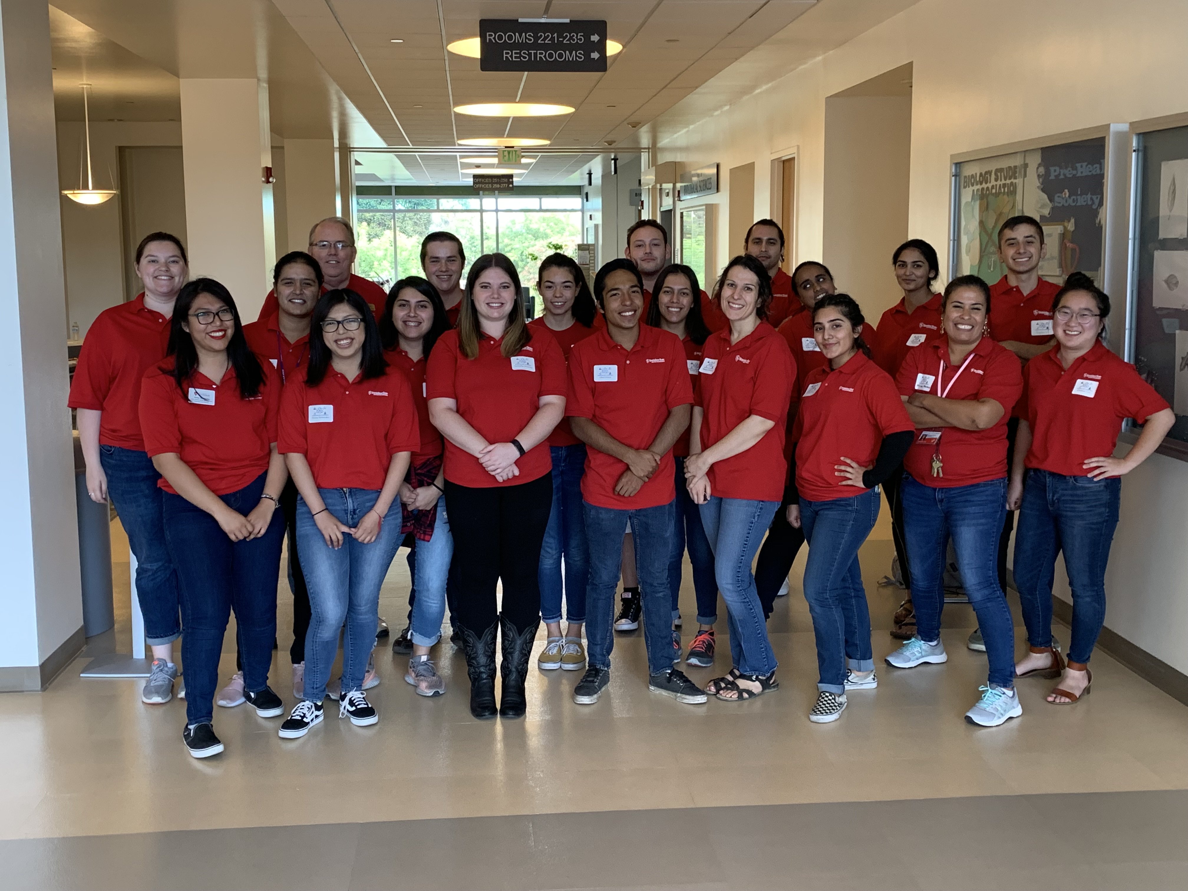 Group of students and staff wearing matching red shirts pose together in a campus hallway for a team photo.