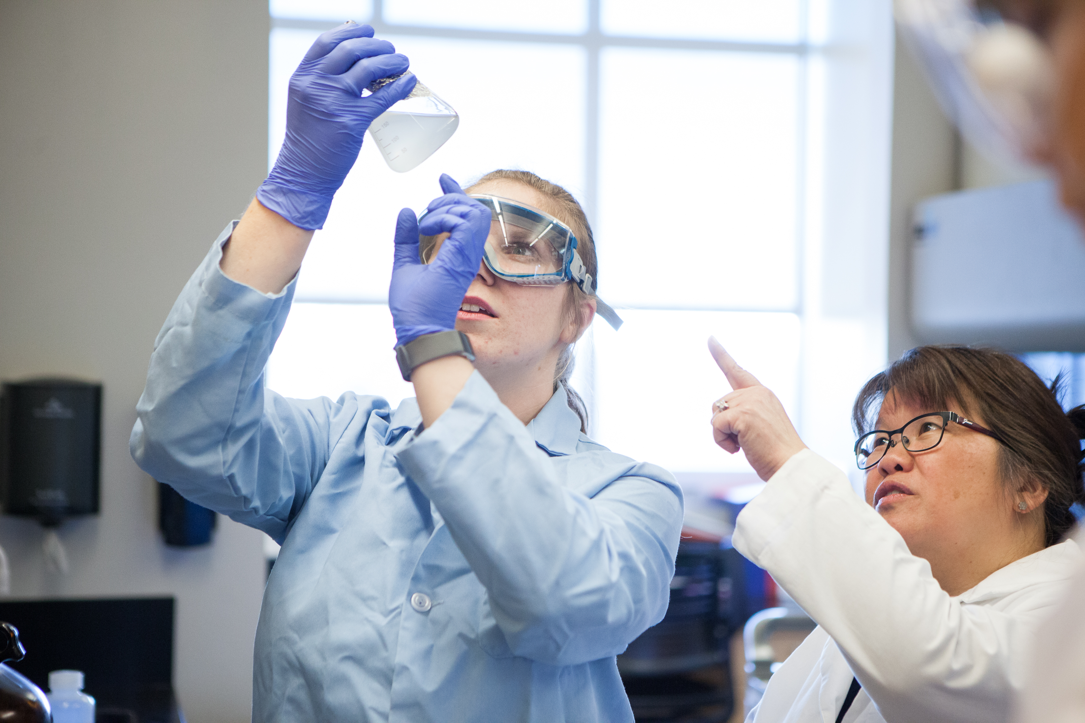 Two scientists in a lab examine a liquid in a flask. One wears goggles and holds it up, while the other points, creating a focused and collaborative atmosphere.