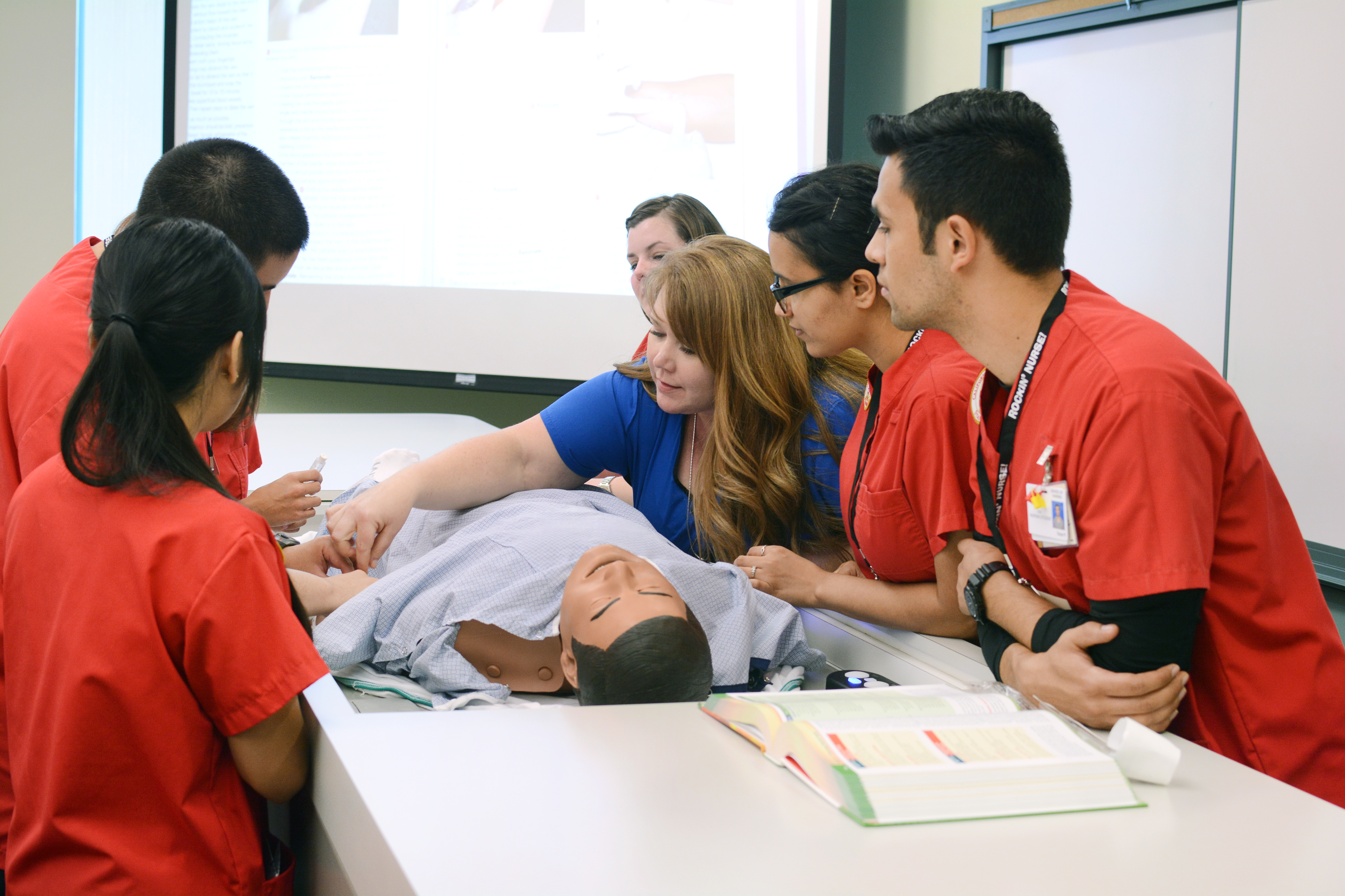 Medical students in red scrubs gather around a mannequin for a hands-on lesson, while an instructor in blue demonstrates techniques. The setting is educational.