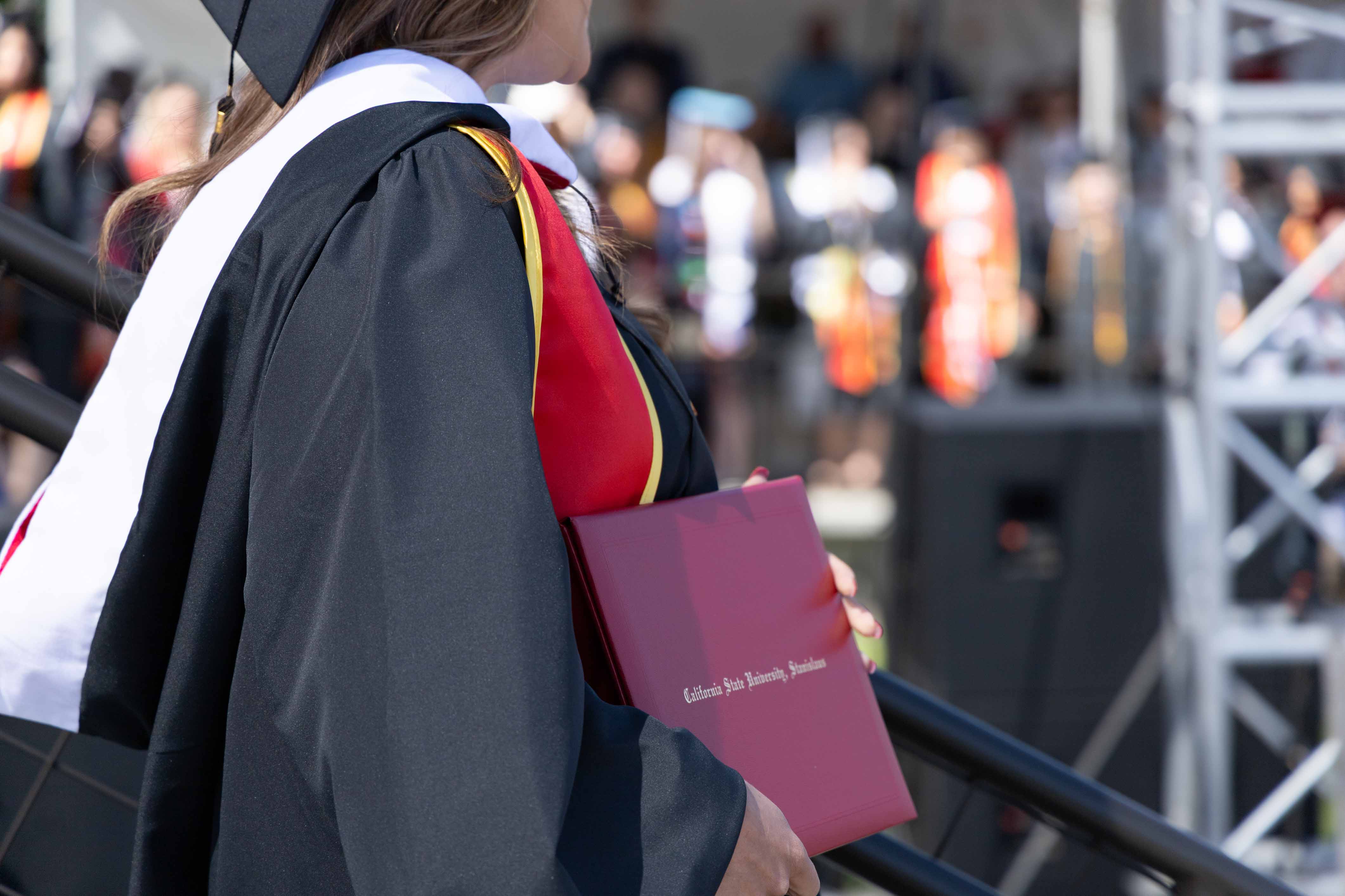 Graduate in a black gown and cap holds a red diploma cover against a blurred background of fellow graduates, conveying achievement and celebration.