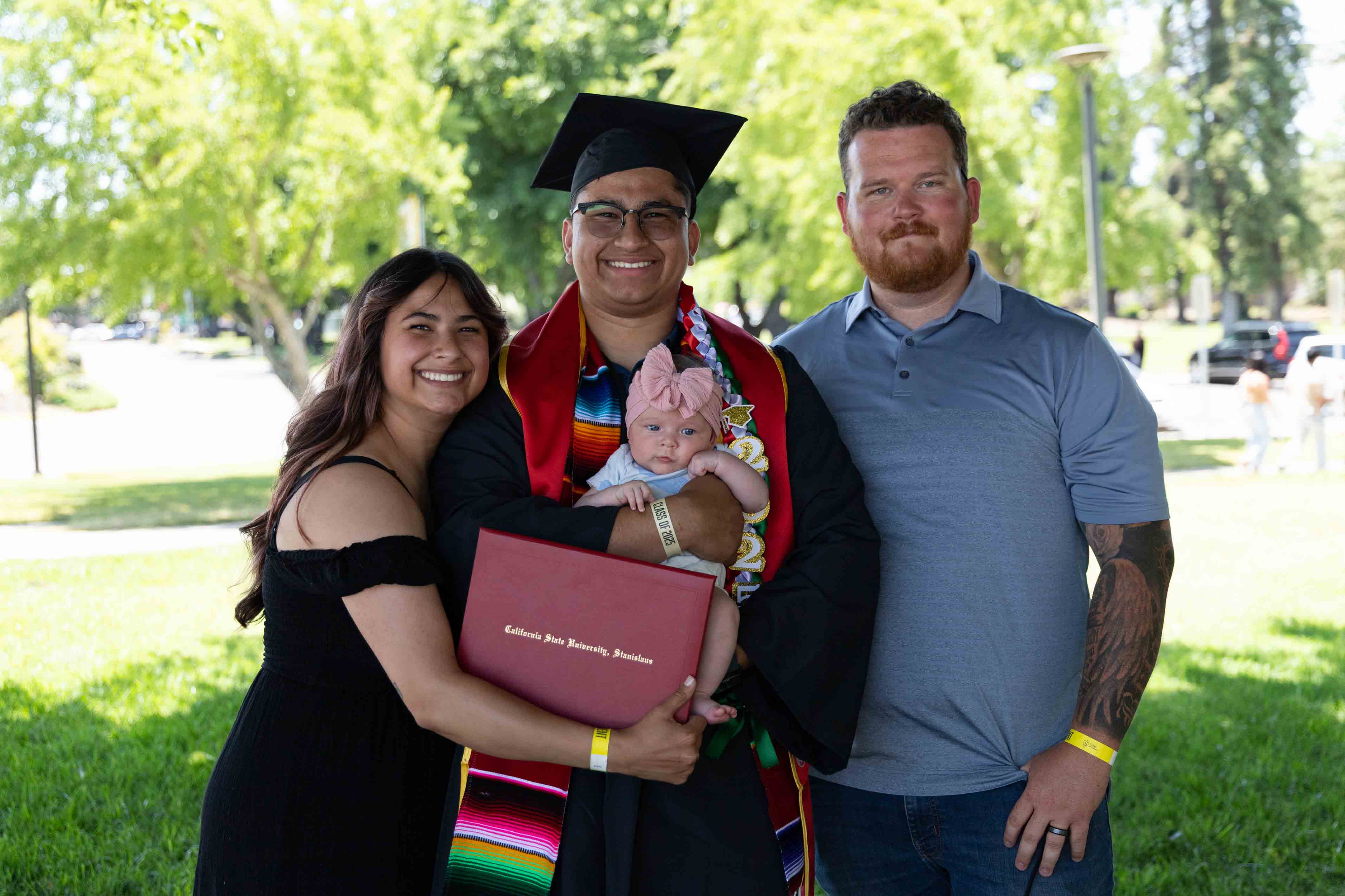 Graduate in cap and gown holds a diploma and baby, flanked by two smiling adults. They stand on a sunny day with trees in the background.