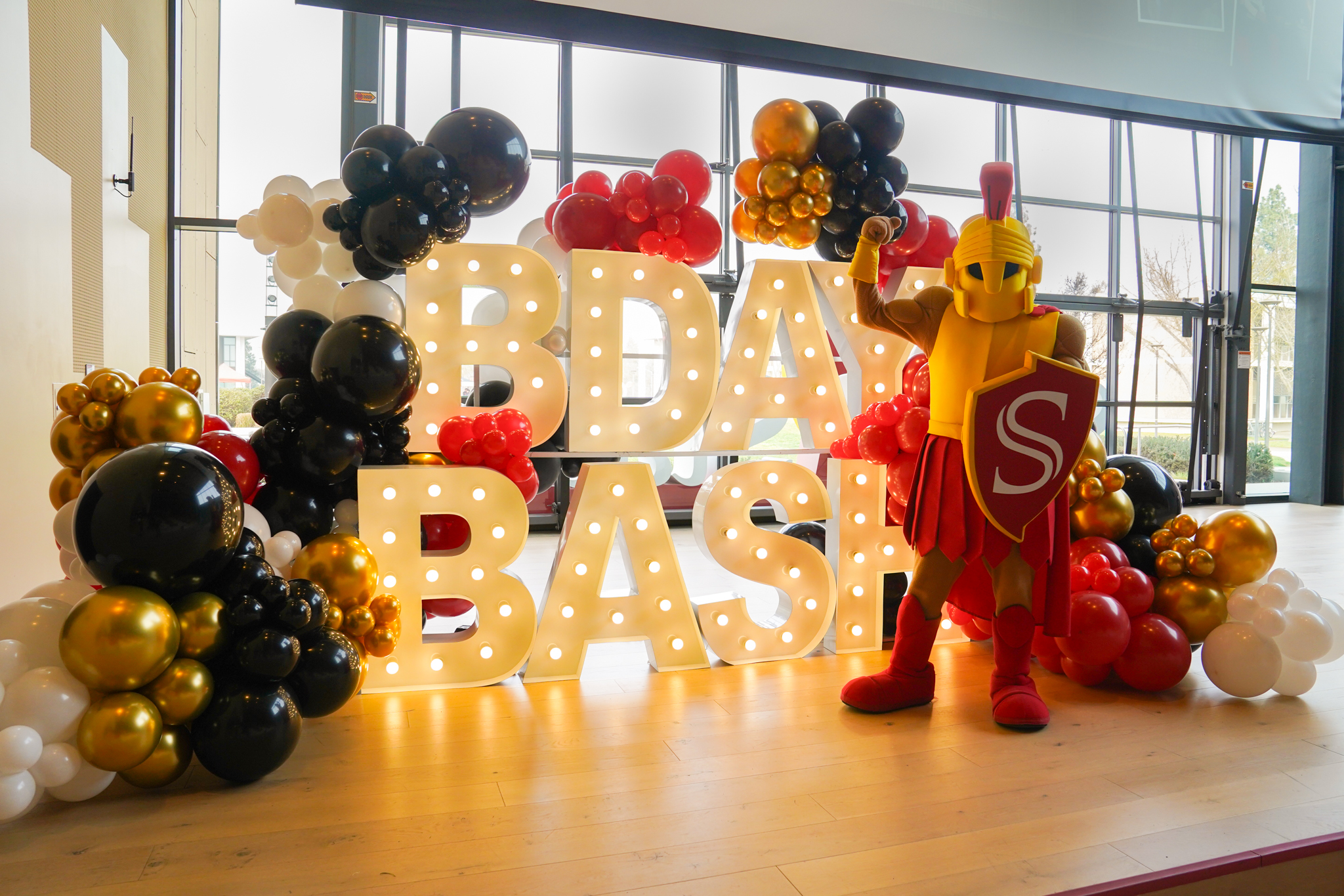 Titus the warrior mascot posing in front of the SC Birthday Bash Sign and Balloons