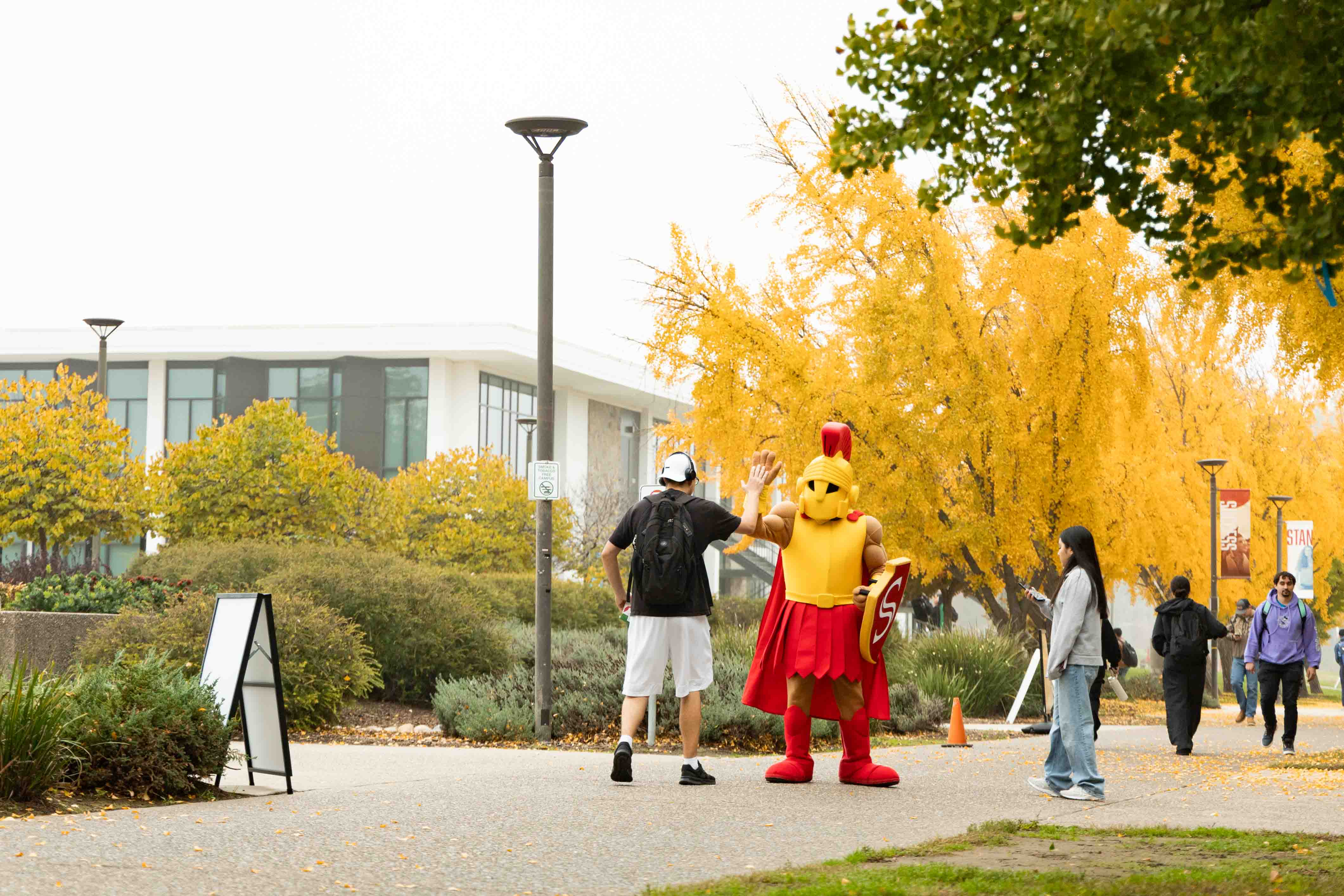 A person high-fives a mascot in yellow and red armor on a tree-lined campus walkway, surrounded by others. Autumn leaves create a vibrant backdrop.