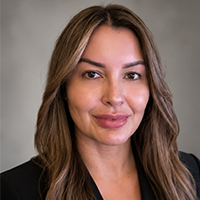 Portrait of a woman with long brown hair, wearing a black top and blazer, smiling slightly against a gray background, conveying professionalism.