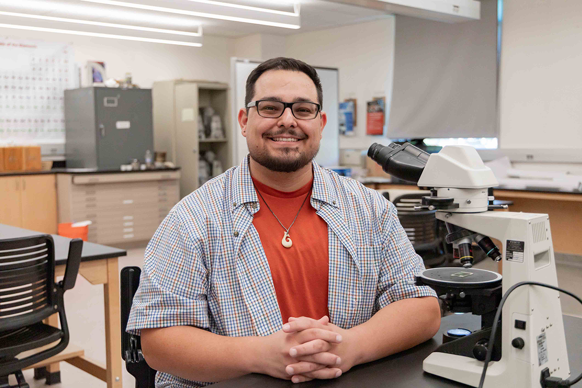 A man with glasses and a beard, smiling, sits at a lab table with a microscope. He wears a red shirt and checkered jacket, conveying a friendly atmosphere.