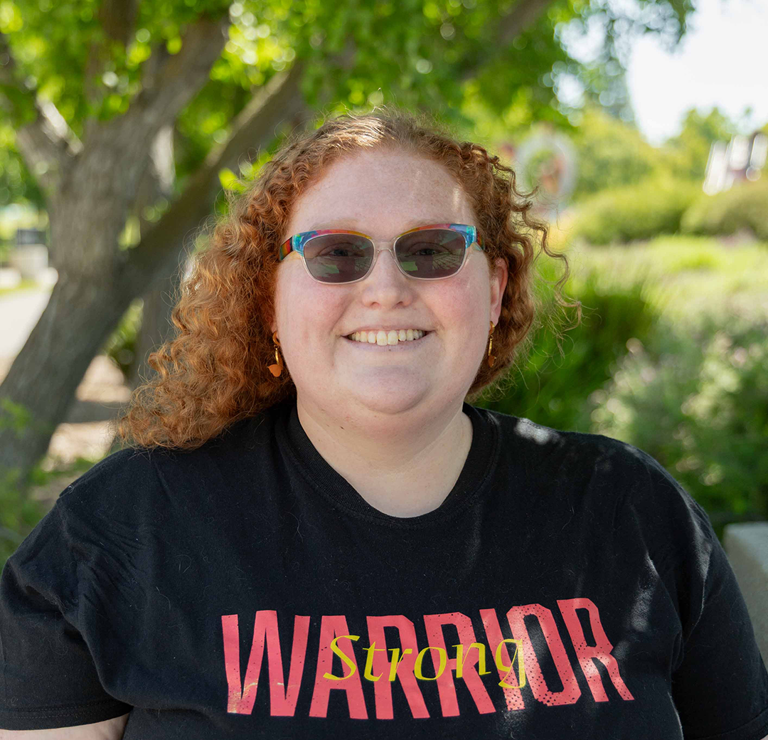 Smiling person with curly red hair and sunglasses wears a black shirt reading "WARRIOR Strong" in red. Trees and greenery form the sunny background.