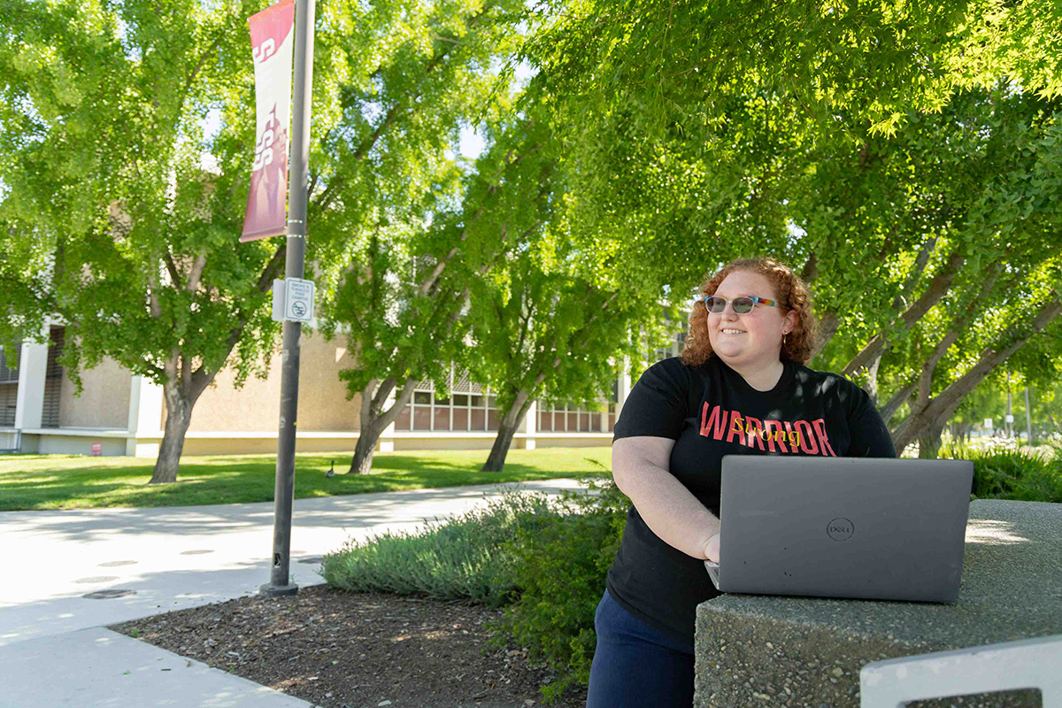 A person with curly red hair, wearing sunglasses and a "Warrior" T-shirt, stands outdoors with a laptop. Green trees and a building are in the background.