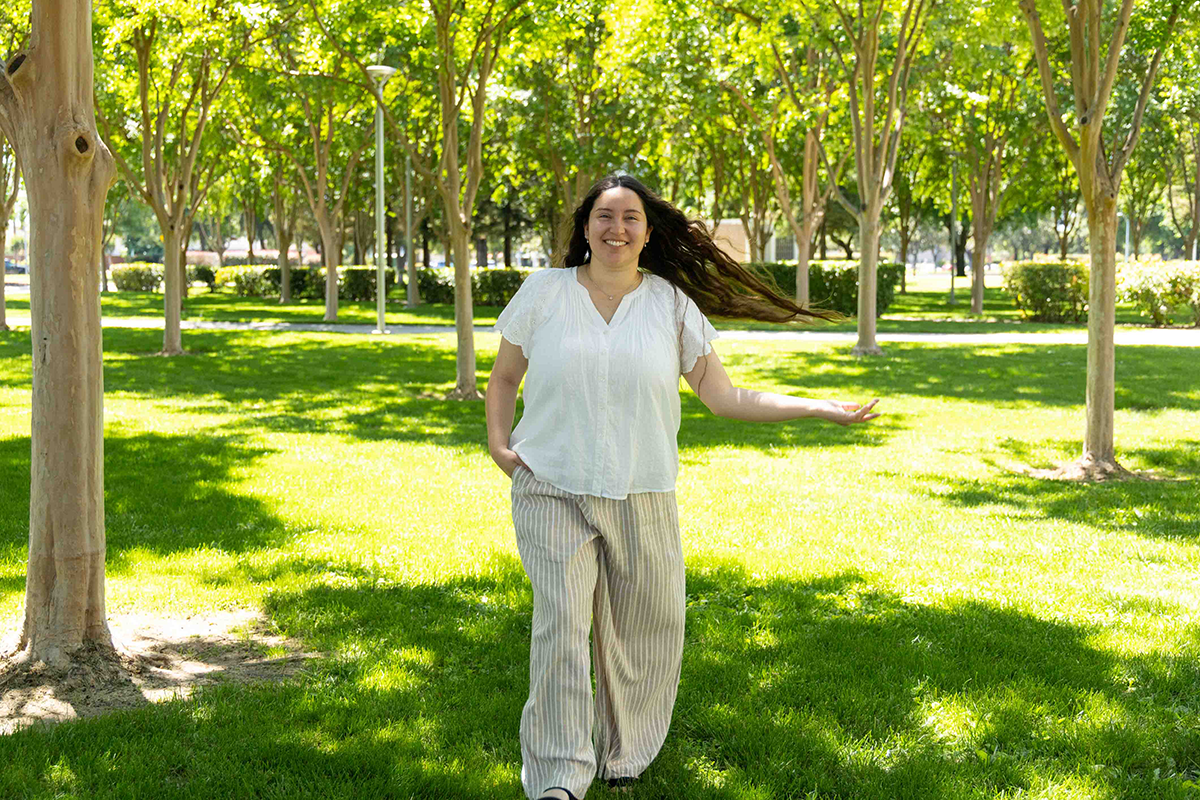 A woman in a white blouse and striped pants joyfully walks through a sunlit park, surrounded by trees and lush grass, exuding happiness and serenity.