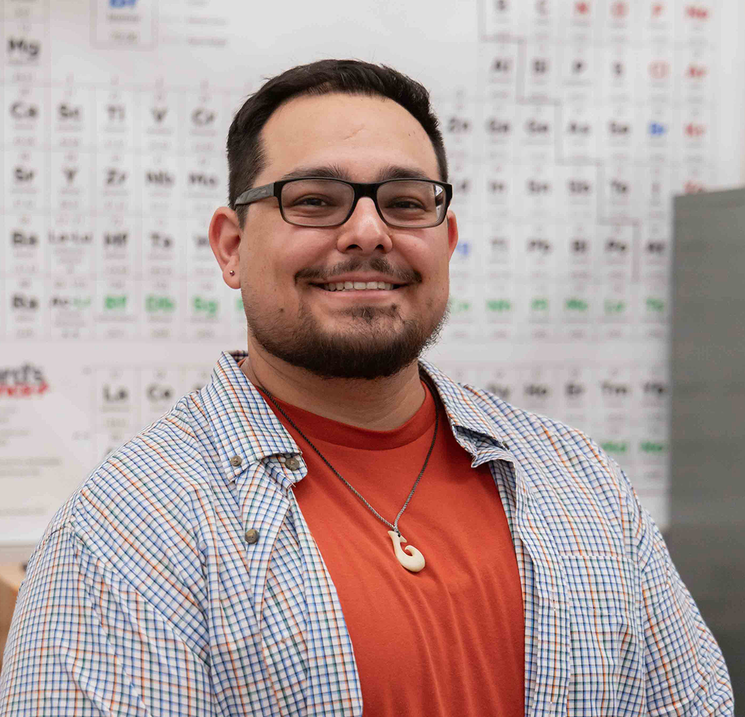 A man with glasses and a beard, smiling, sits at a lab table with a microscope. He wears a red shirt and checkered jacket, conveying a friendly atmosphere.