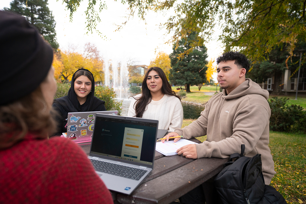 Four students sit around an outdoor picnic table, talking and working with laptops and notebooks near a campus fountain.