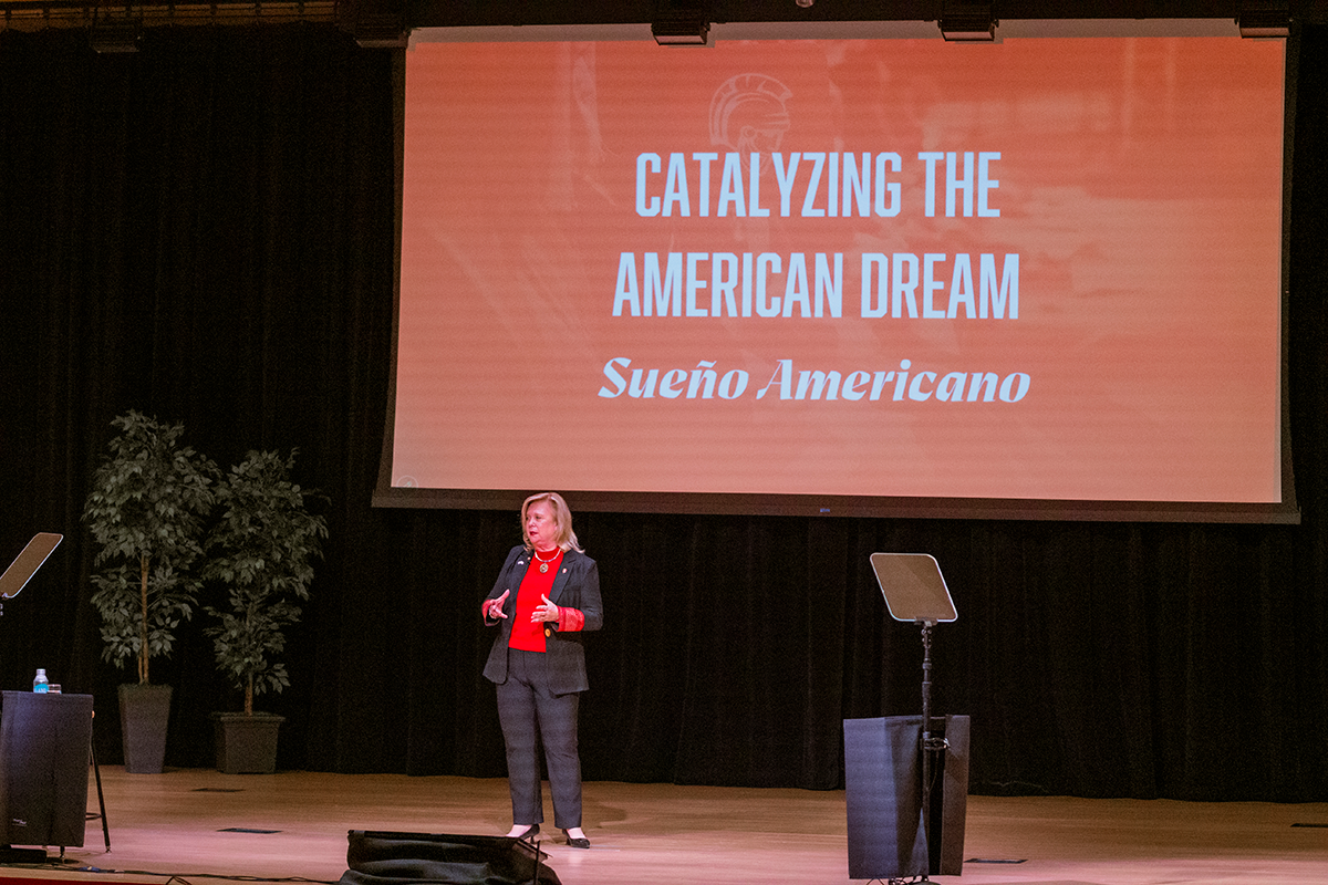 President Britt Rios-Ellis speaks onstage beneath a screen displaying “Catalyzing the American Dream” and “Sueño Americano.”