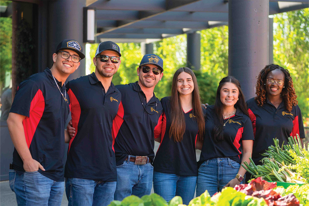 Six smiling people wearing matching black-and-red Stan State Agribusiness shirts pose behind a display of fresh vegetables.