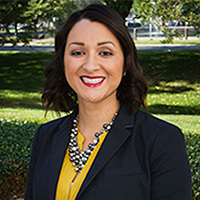 Professional headshot of a smiling woman in a black blazer and yellow blouse, wearing layered necklaces, posed outdoors in a sunny park.