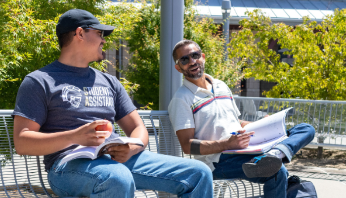 Two students talking on a campus bench with notebooks and backpacks. 