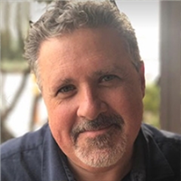 Close-up portrait of a friendly middle-aged man with salt-and-pepper hair and goatee, wearing a dark shirt near a window.