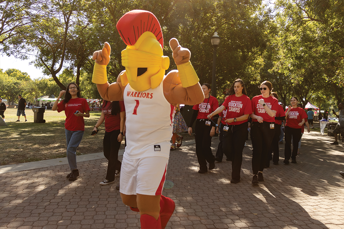 Stanislaus State mascot Titus gives two thumbs up while walking with students and staff during an outdoor campus event.