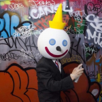  a person dressed in a suit and wearing a Jack in the Box fast food company mask holds a spray can against a wall with grafitti