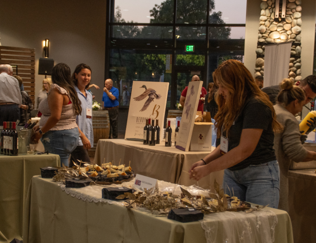 Individuals enjoying a social event, standing around tables adorned with wine glasses and various food items.