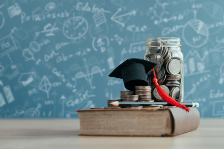 a book with change, pencils and a graduation cap stacked on top. With a chalkboard in the background.