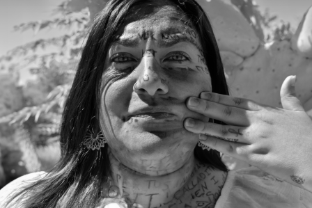 Black and white photograph depicting a woman focused on the camera. It's titled Calladita Te Ves Más Bonita by Artist Monica Crystal Ocegueda. 