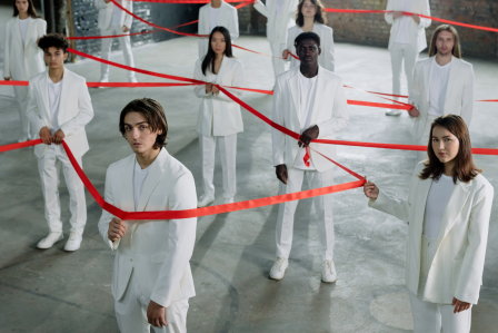 Various people stand in a room holding tape in a pattern that shows connectedness.