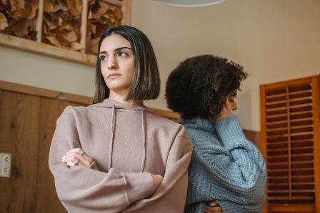 Two women stand back-to-back indoors, appearing upset.