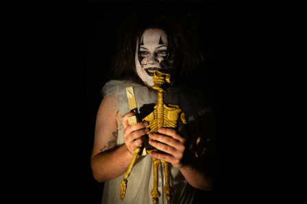 Someone with black and white clown makeup holds and plays with a skeleton toy