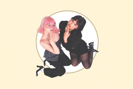 Two people kneeling, one with pink hair, glasses, and a playful expression, the other with dark hair and black outfit, both posing thoughtfully.