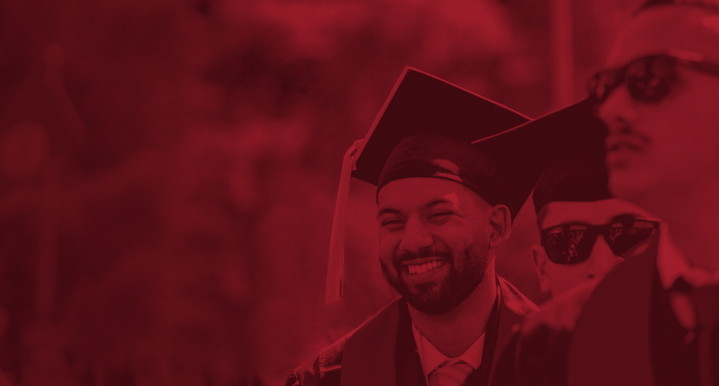 Two graduates in caps and gowns, one wearing sunglasses, celebrate during a ceremony in a vibrant red-toned background.