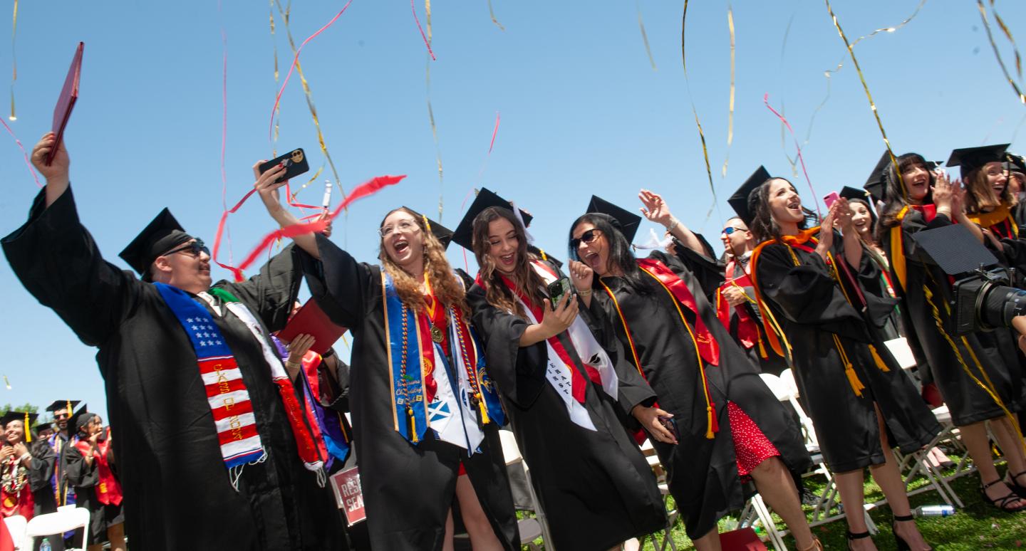 Graduates in caps and gowns celebrate joyfully under a clear sky, with colorful streamers in the air and diplomas held high, capturing excitement.