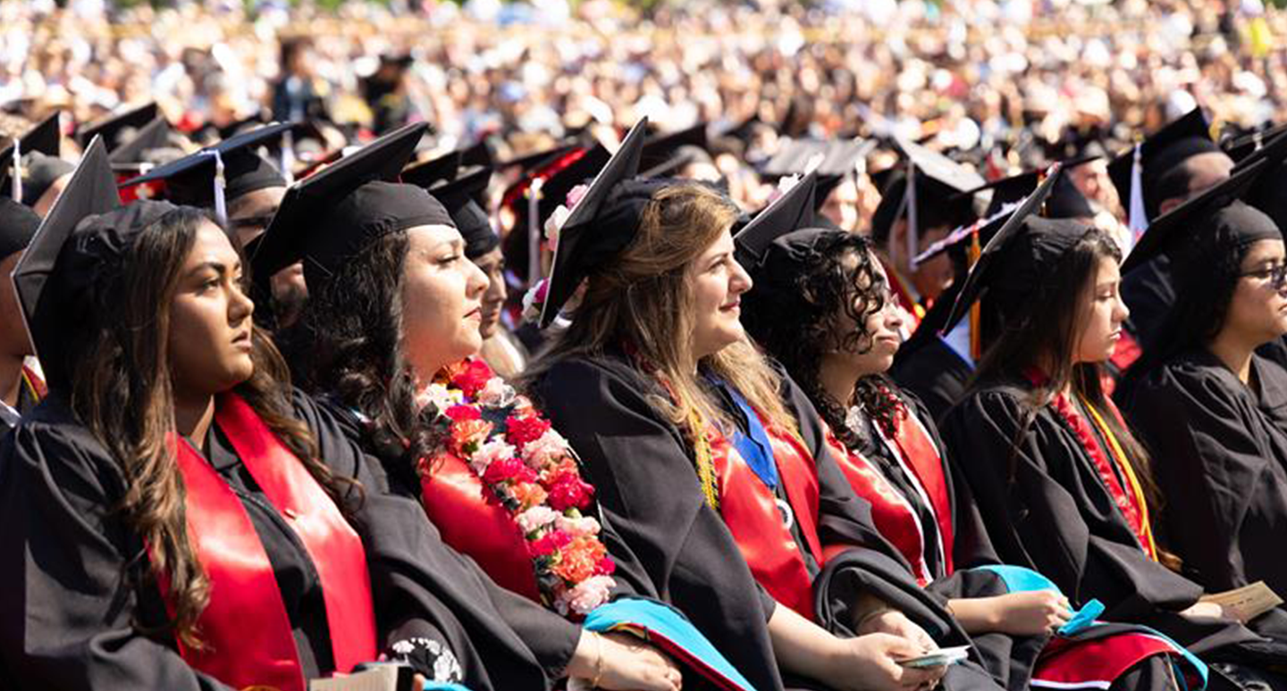 A crowd of graduates in black caps and gowns sits attentively, adorned with sashes and leis, at a ceremonial outdoor event.