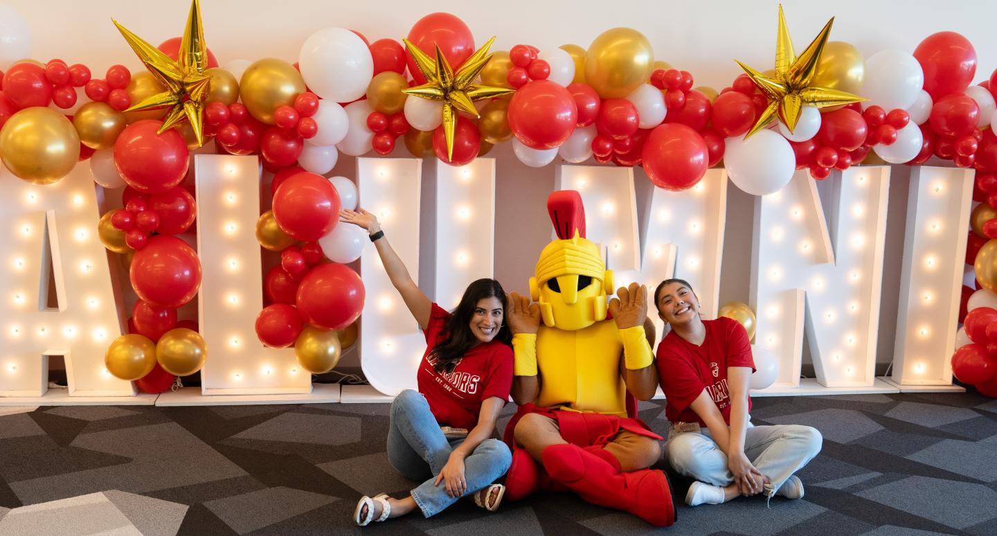 Two people and a mascot sit happily on a patterned floor in front of large illuminated "ALUMNI" letters. Red, white, and gold balloons with star shapes decorate above.