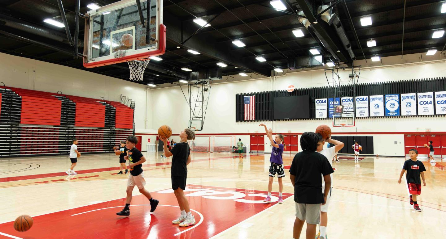 Children practice basketball in a gym with red and black bleachers. An American flag and championship banners hang on the wall above them.