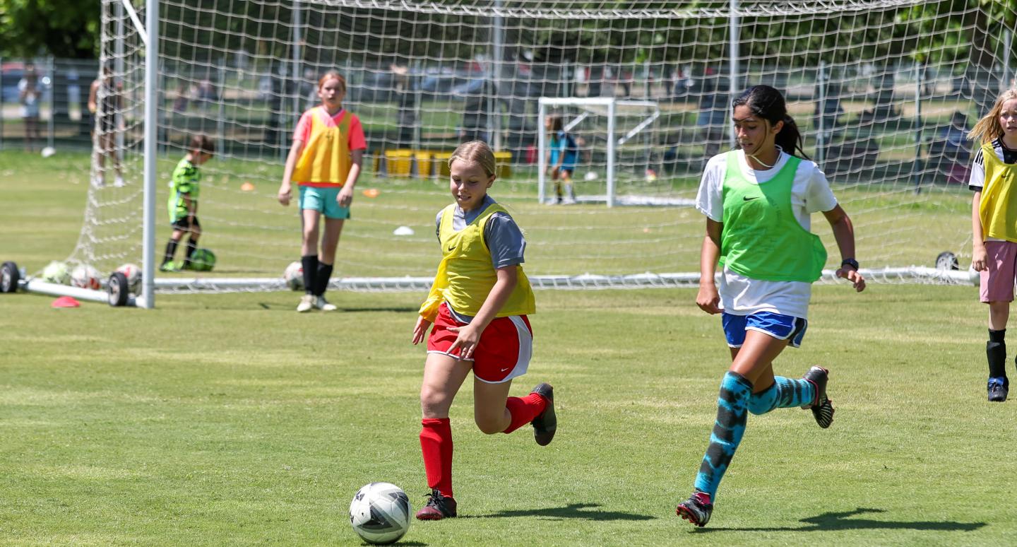 Two girls compete for a soccer ball on a sunny field. A girl in a yellow bib dribbles while another in green chases. Others watch near the goal.