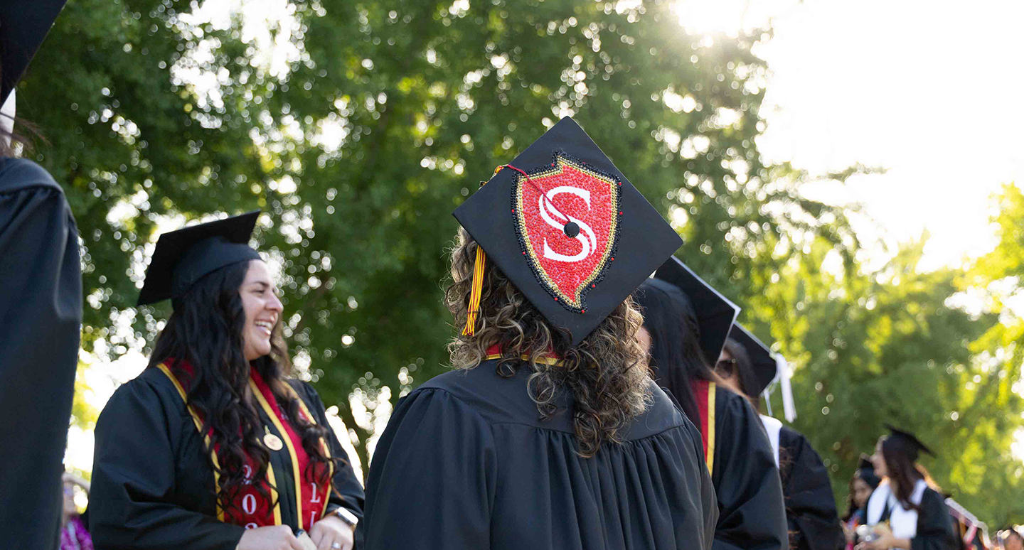 Graduates in caps and gowns gather outdoors, celebrating. One cap is adorned with a decorative red "S" emblem. Sunlight filters through trees, creating a joyful atmosphere.