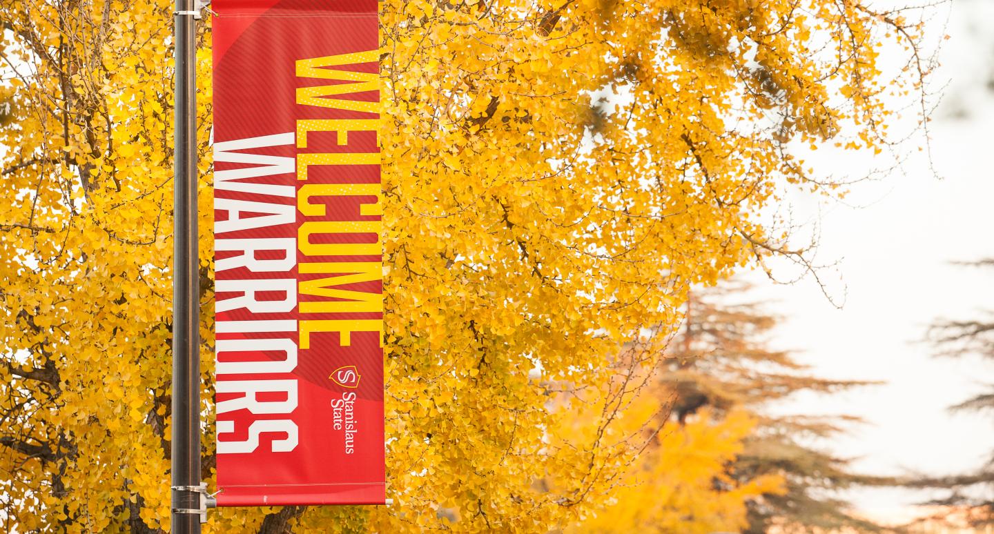A red vertical banner reading "Welcome Warriors" hangs on a pole, surrounded by vibrant yellow autumn leaves. The tone is welcoming and festive.