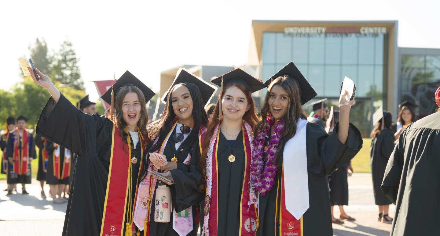 Four graduates in caps and gowns, smiling joyfully, pose together outdoors. They exude happiness and pride, holding diplomas before a university building.