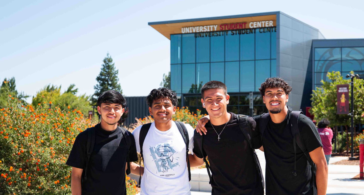 Four smiling young people stand arm in arm in front of a modern university student center, surrounded by bright orange flowers and greenery, conveying a sense of camaraderie and positivity.